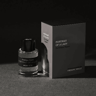 Minimalist product shot showing the Frederic Malle Portrait Of A Lady travel spray nestled in its box, beside the larger full-size fragrance packaging in the background.